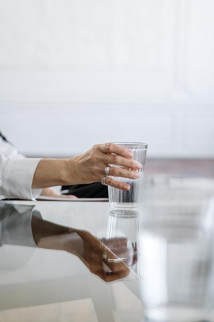 approach-img Close-up of a hand with rings holding a glass on a glass table, perfect for health and lifestyle themes.